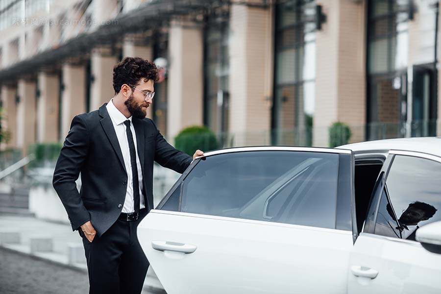 年轻英俊的成功经理穿着黑色西装进入他的汽车后座，靠近现代商务中心，在大城市的街道上