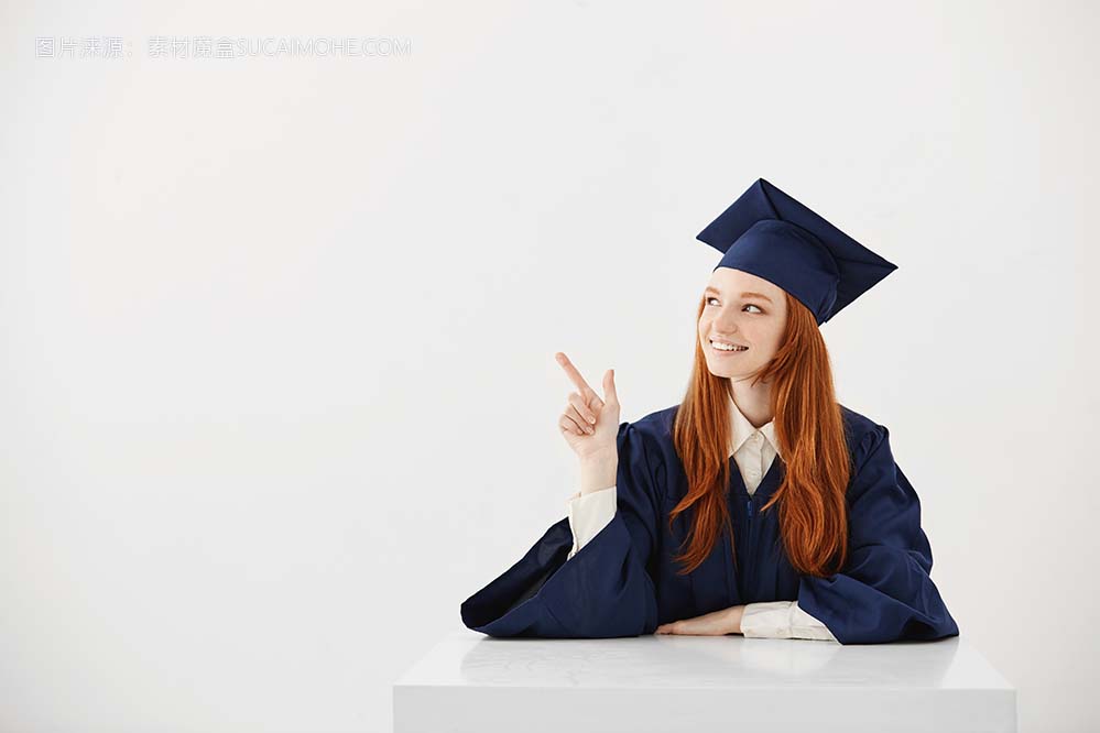 年轻的女大学毕业生在学术帽坐在桌前微笑着指着左边。未来的律师或工程师展示一个想法